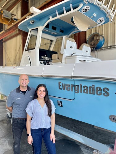 A man (left) standing next to a woman (right). Both are smiling and posing in front of a boat that is on a boat trailer.