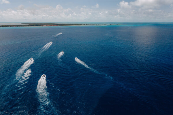 Aerial view of multiple Everglades boats speeding through the open ocean leaving white wakes behind them with a distant shoreline and cloudy sky in the background