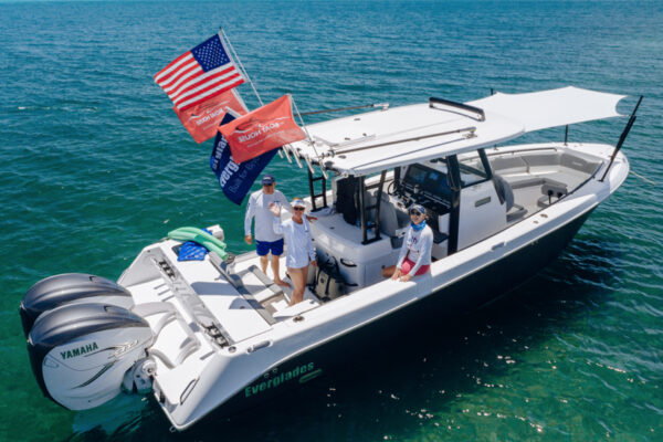 An Everglades boat floating on clear water with three people on board waving, multiple flags flying from the back, and two Yamaha engines at the stern