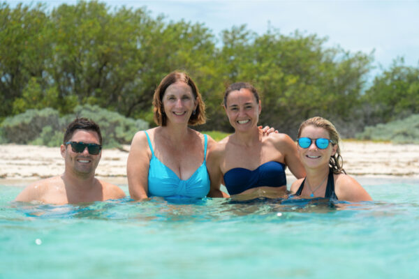 Four people smiling and standing in shallow turquoise water near a sandy shoreline with green shrubs in the background