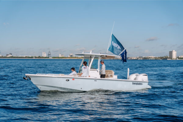An Everglades boat cruising on calm water with three people on board, flying an Everglades flag, and a city skyline visible in the background