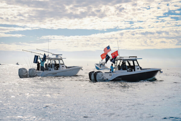 Two Everglades boats floating on calm water with multiple flags flying at the back under a cloudy sky with other boats visible in the distance