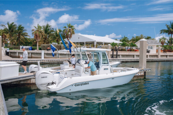 An Everglades boat docked at a marina with people on board preparing for a trip under a sunny sky with palm trees and other boats in the background