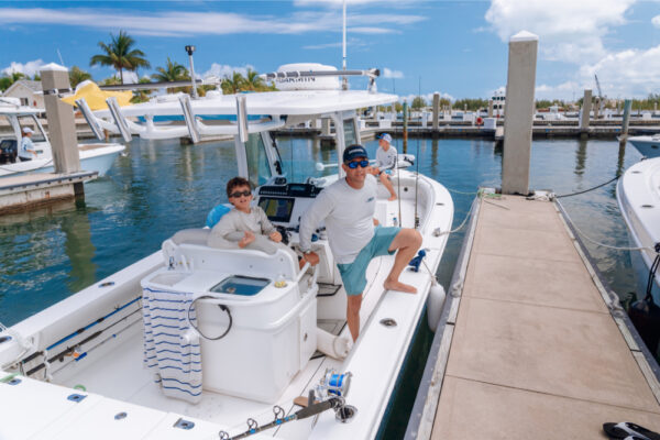 Two people sitting on an Everglades boat docked at a marina on a sunny day with other boats and palm trees in the background