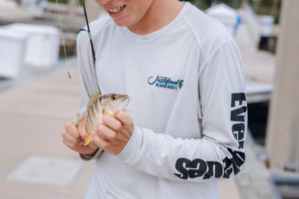 A person holding a small fish they just caught while wearing a long sleeve Everglades shirt at a marina