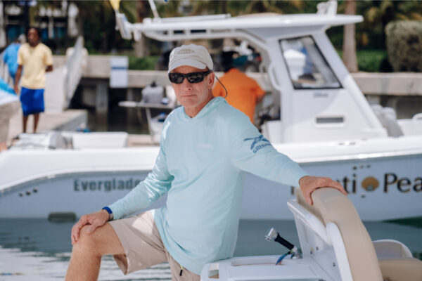 A man wearing sunglasses and a light blue shirt leaning on an Everglades boat at a marina with another Everglades boat in the background