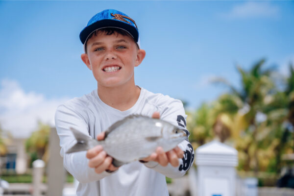 A young boy smiling and holding a small fish in his hands, wearing a long-sleeve shirt and a blue cap, with a sunny marina and palm trees in the background