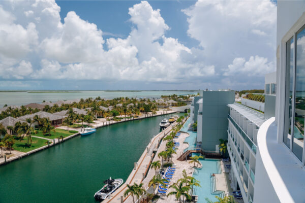 Aerial view of a marina with boats docked along a waterway surrounded by palm trees and buildings with a large hotel and pool area on the right side under a partly cloudy sky