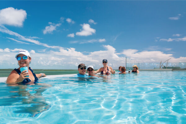 A group of people relaxing in a pool on a sunny day with clear blue skies and scattered clouds wearing hats and sunglasses while enjoying drinks