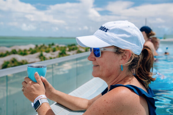 A woman wearing an Everglades hat and sunglasses relaxing in a pool while holding a drink with a scenic view of palm trees and the ocean in the background under a partly cloudy sky