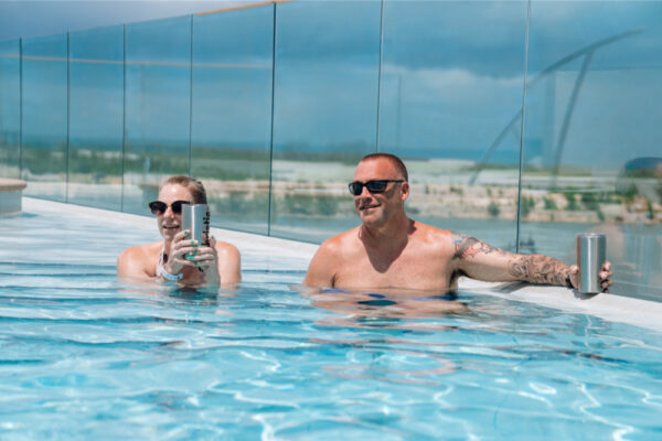 Two people relaxing in a pool holding stainless steel tumblers and wearing sunglasses with a glass fence and a landscape visible in the background