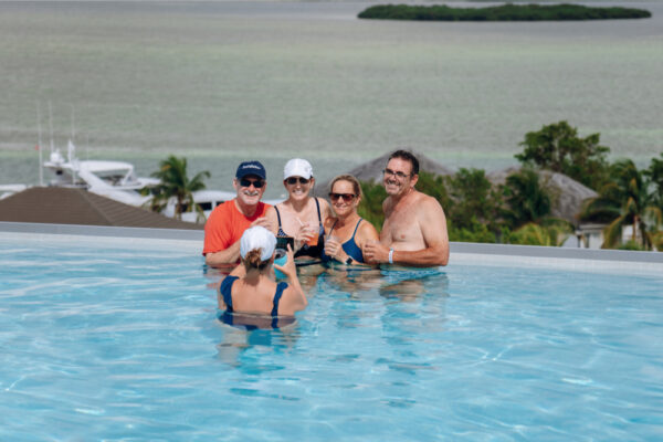 A group of five people standing in an infinity pool overlooking a scenic view of the ocean and trees, smiling and enjoying drinks together
