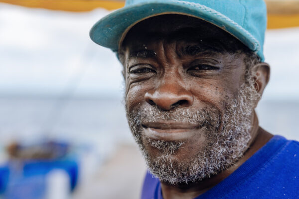 A close-up of a man with a beard wearing a blue hat and shirt, smiling slightly, with a blurred background of blue and yellow tones