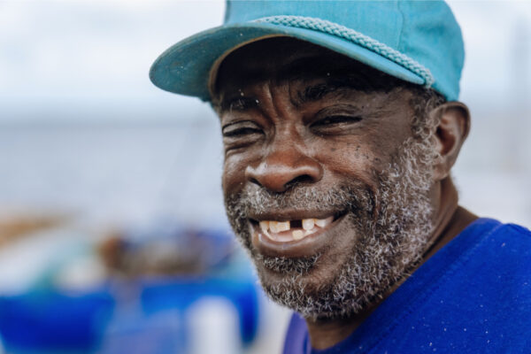 A close up of a smiling man wearing a blue hat with a blurred background of blue and white tones