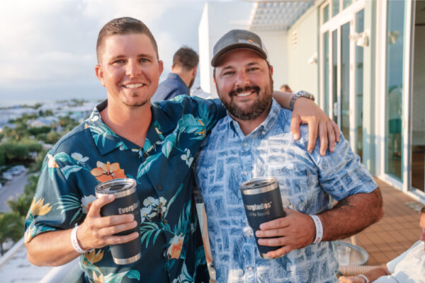 Two men smiling and standing together on a deck, each holding an Everglades-branded tumbler, wearing casual floral shirts during a gathering