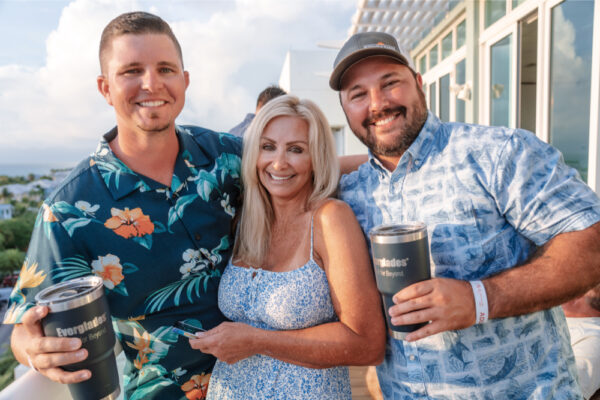 Two men and a woman smiling together on a deck, with the men holding Everglades-branded tumblers, wearing casual floral shirts during a gathering