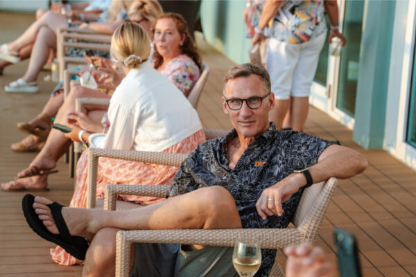A man sitting in a chair on an outdoor deck wearing glasses and a patterned shirt with other people seated and talking in the background