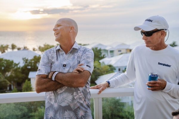Two men standing on a balcony at sunset with one man smiling and the other holding a drink while wearing an Everglades shirt and sunglasses with a scenic view of rooftops and trees in the background