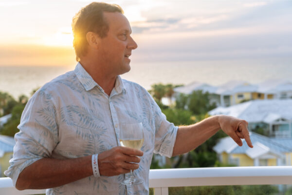 A man holding a glass of wine while standing on a balcony at sunset pointing and talking with a scenic view of rooftops and the ocean in the background