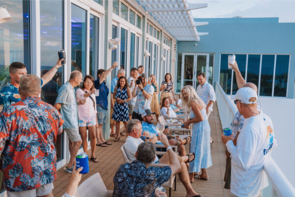 A group of people gathered on a large outdoor deck raising drinks and toasting with some seated and others standing during a casual event at a building with large windows