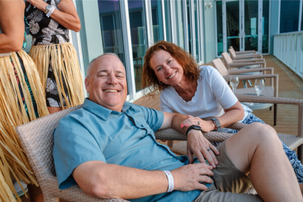 A man and woman sitting and smiling on an outdoor deck during a casual gathering with other people standing in the background wearing grass skirts