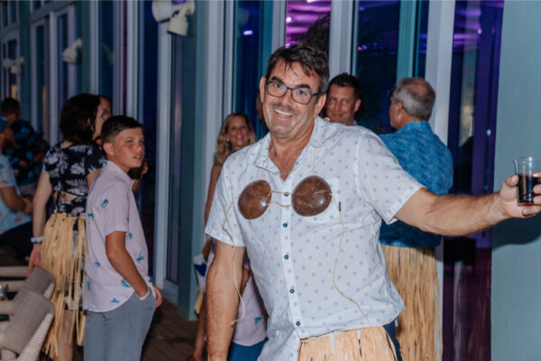 A man smiling and posing for the camera at a party wearing a coconut bra and grass skirt with other people in the background