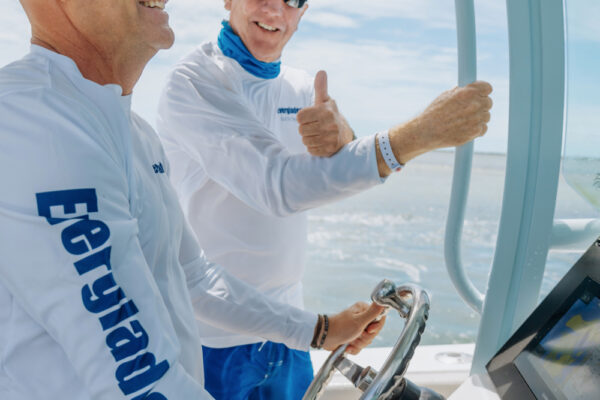 Two men wearing sunglasses and white shirts steering a boat, with one man giving a thumbs up gesture