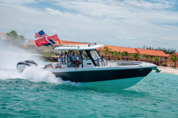An Everglades boat speeding across turquoise water with multiple flags flying at the back and a shoreline with orange roofed buildings and palm trees in the background