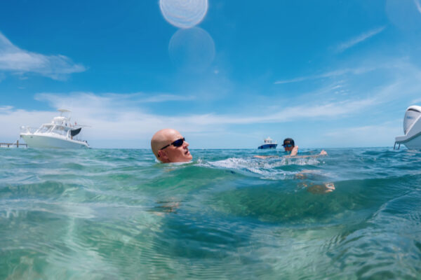 Two people swimming in clear turquoise water with Everglades boats in the background under a bright blue sky
