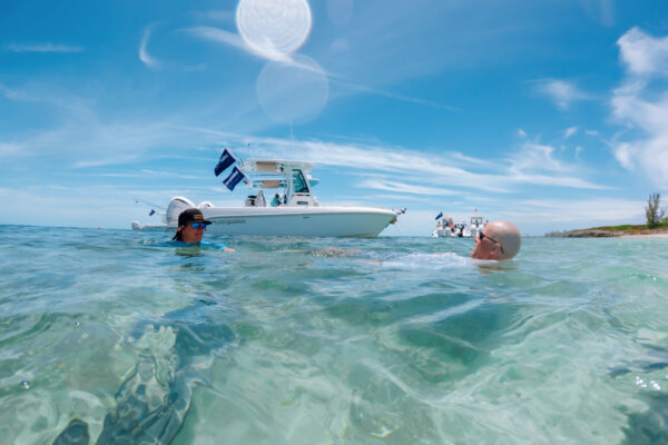 Two people swimming in clear turquoise water with an Everglades boat floating nearby under a bright blue sky