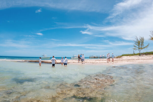 A group of people wading in shallow water near a sandy beach with boats docked in the distance under a bright blue sky